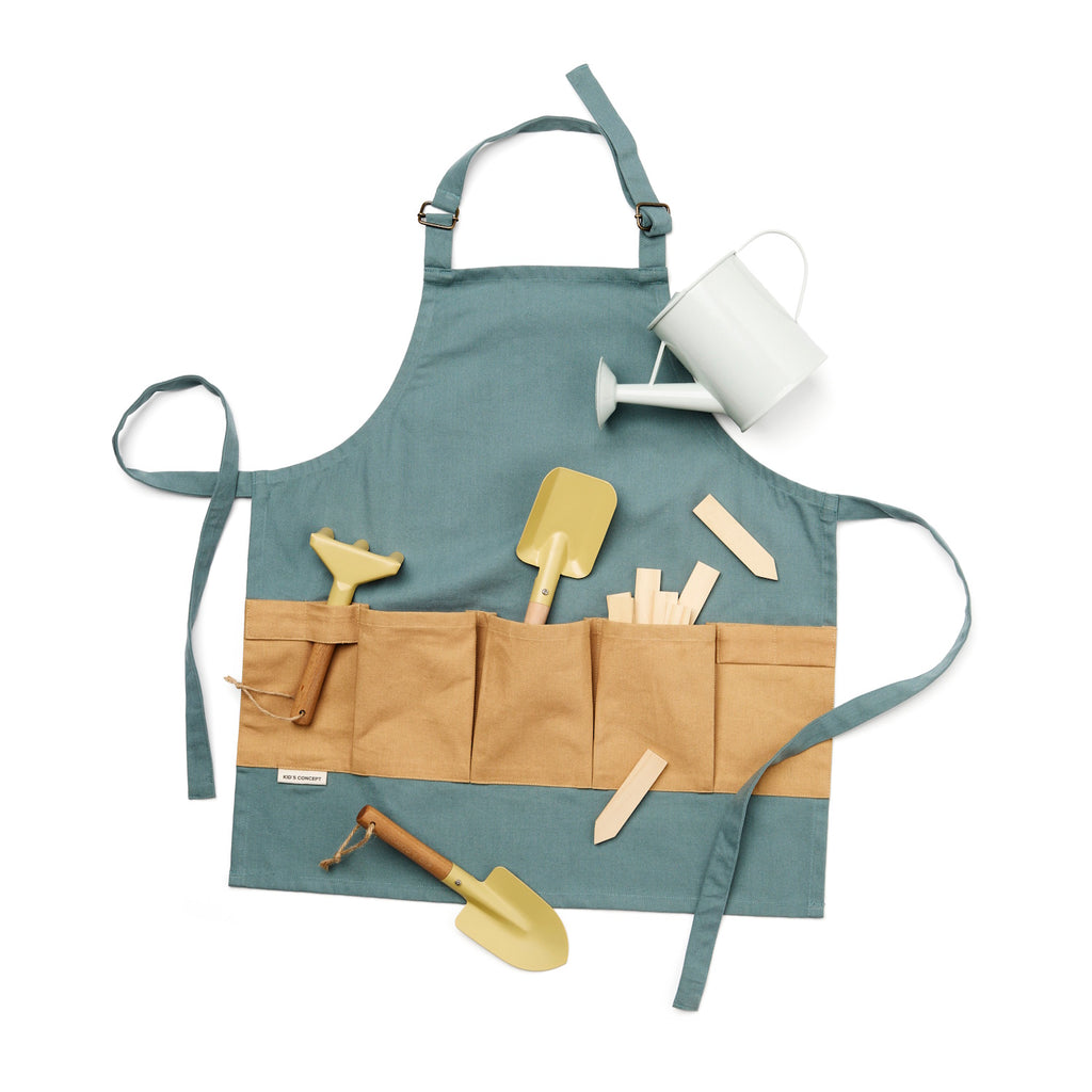 A teal and tan gardening apron holds a white watering can, small shovel, spade, rake, and wooden plant markers. The setup suggests a gardening pretend play.