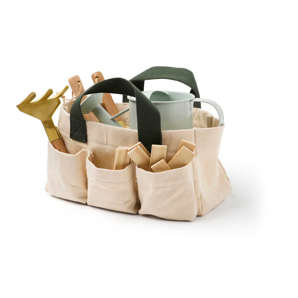 A beige canvas gardening tote with dark green handles holds a metal watering can, wooden stakes, and a small rake. The scene conveys readiness and organization.