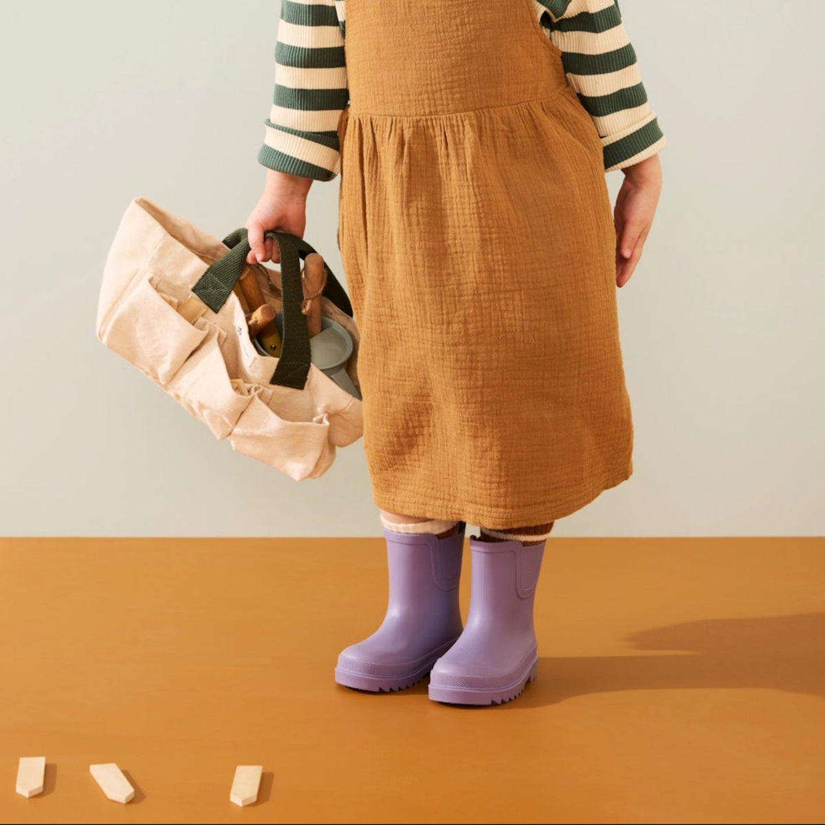 Young child in brown pinafore dress holding a canvas garden tote bag with toy tools inside.