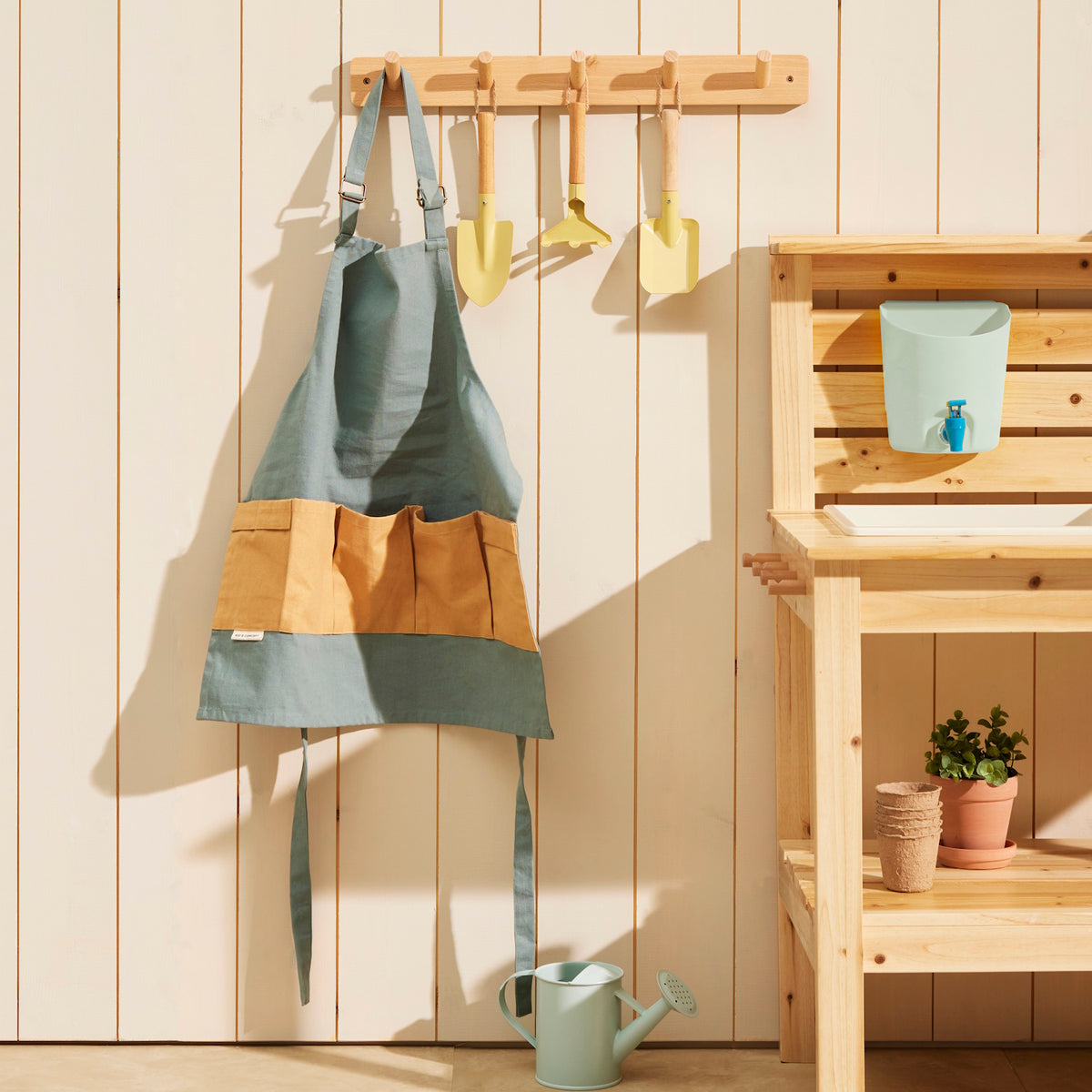 Green kids gardening apron and small yellow tools hanging on a wooden wall rack next to a play bench.