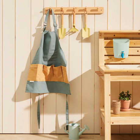 Green kids gardening apron and small yellow tools hanging on a wooden wall rack next to a play bench.