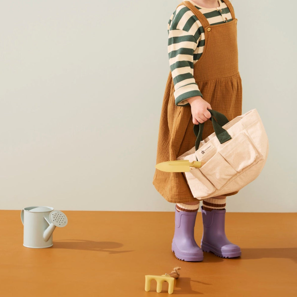 Young child wearing purple boots holding a canvas garden tote next to a toy watering can and rake.