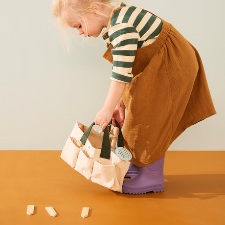 Young child in purple boots leaning down to set a canvas tool bag filled with toy garden supplies.