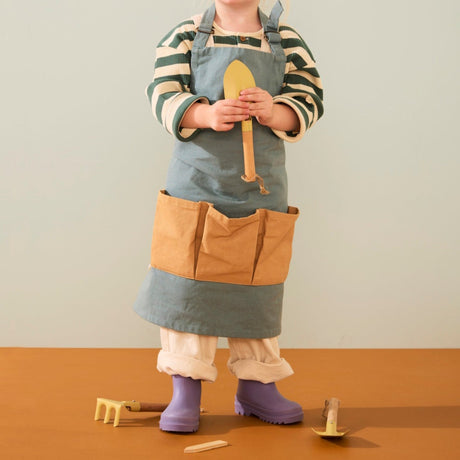 Young child wearing a green apron holding a toy trowel with gardening tools at their feet.