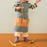 Young child wearing a green apron standing and examining a small yellow toy rake.