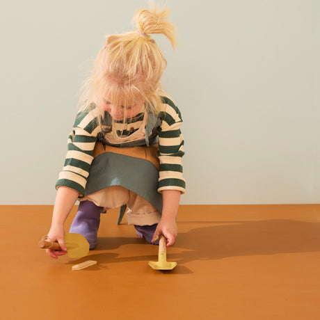 Young child in a green apron crouching down to play with small yellow toy garden tools.