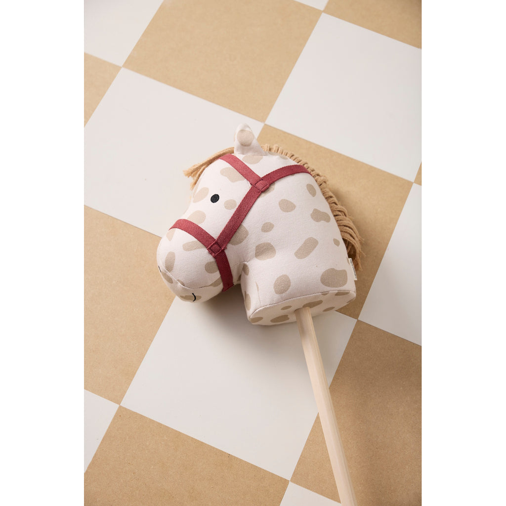 A playful rocking horse head on a stick with a cream base and brown spots. It features a red bridle and yarn mane on a simple wooden stick.