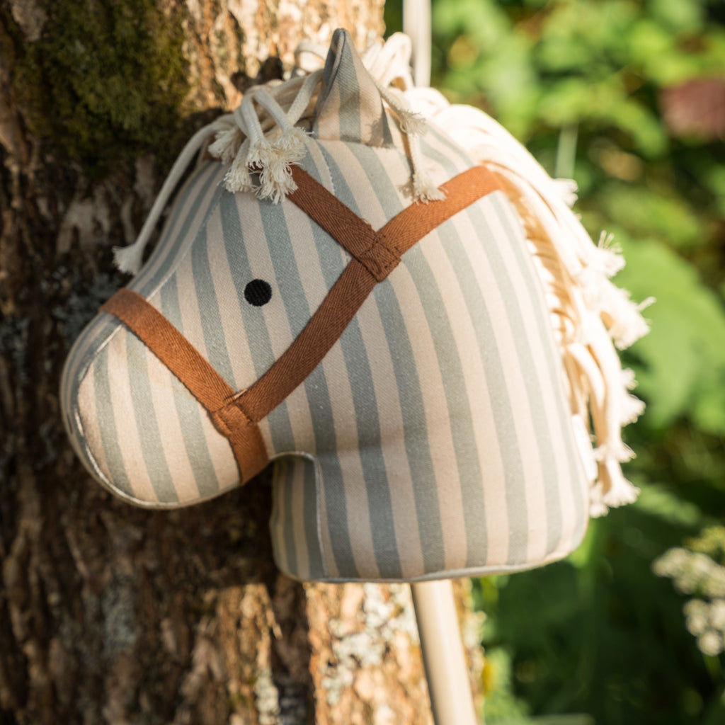 Striped fabric hobby horse toy with a beige and light blue color scheme, featuring a simple face and yarn mane, attached to a wooden stick.