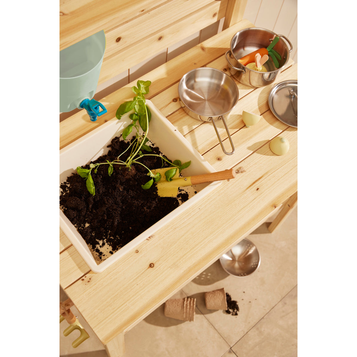 Top-down view of a wooden play mud kitchen counter featuring a sink bin filled with potting soil, a plant, a small shovel, and metal play cookware.