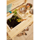 Top-down view of a wooden play mud kitchen counter featuring a sink bin filled with potting soil, a plant, a small shovel, and metal play cookware.
