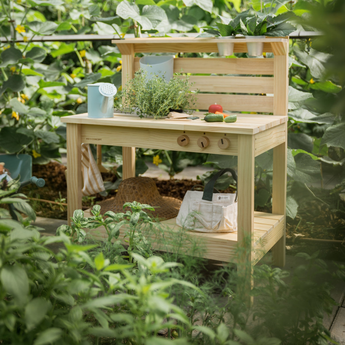 Wooden play mud kitchen set up in a lush garden greenhouse surrounded by plants.
