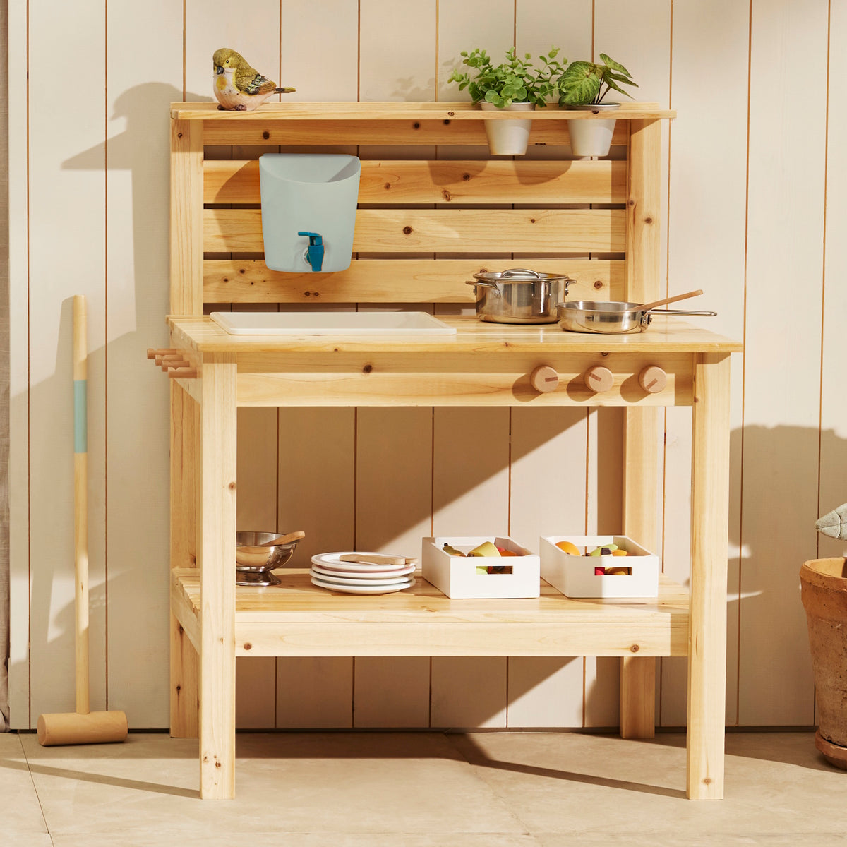 Wooden play mud kitchen arranged with pots, pans, small potted plants, and storage crates against a wooden wall background.