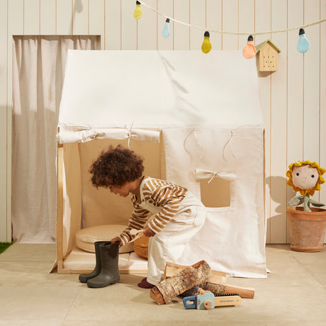Child leaning into an off-white play tent near toy boots, wooden logs, and colorful garland.