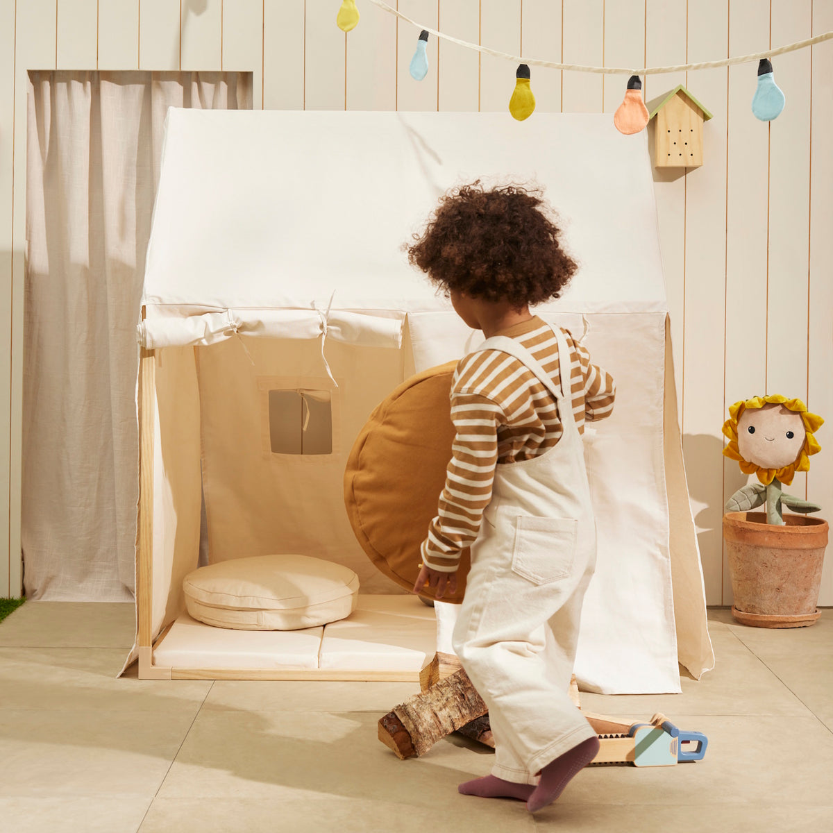 Child carrying a round mustard cushion near an off-white play house tent and floor cushions.