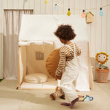 Child carrying a round mustard cushion near an off-white play house tent and floor cushions.