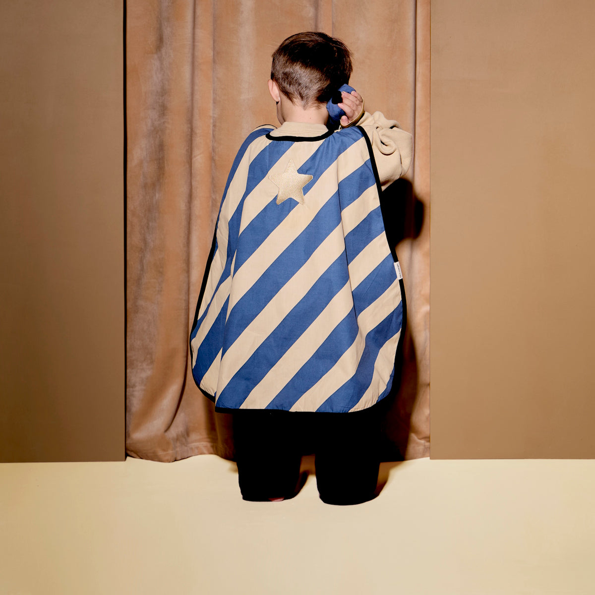 Child from behind wearing a striped dress-up cape, holding a toy walkie-talkie to their ear.
