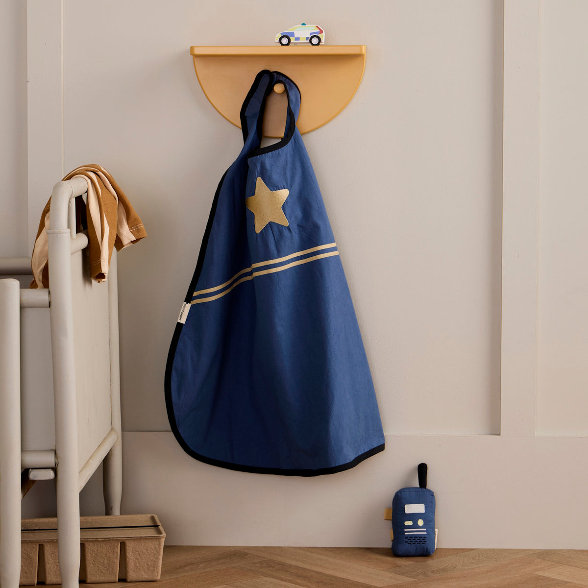 Blue dress-up cape with a gold star badge hanging on a hook next to a toy walkie-talkie.