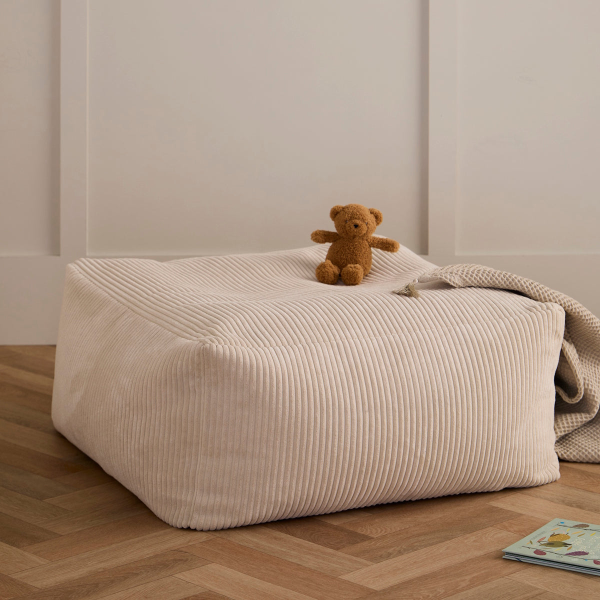 An off-white corduroy floor pouffe with a small teddy bear and blanket resting on top.