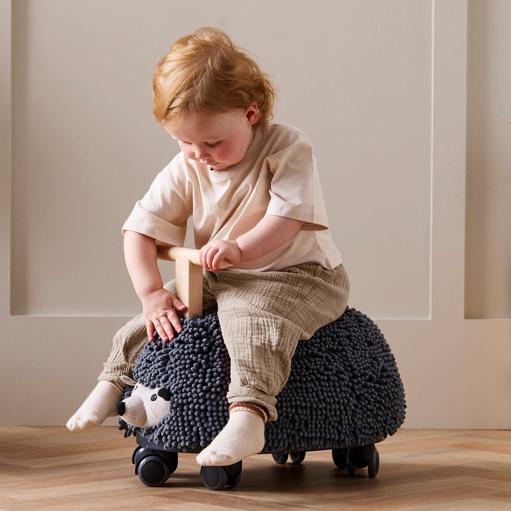Toy hedgehog with a textured, dark gray surface resembling bristles, mounted on four wheels. It has a wooden handle for riding. Playful and whimsical.