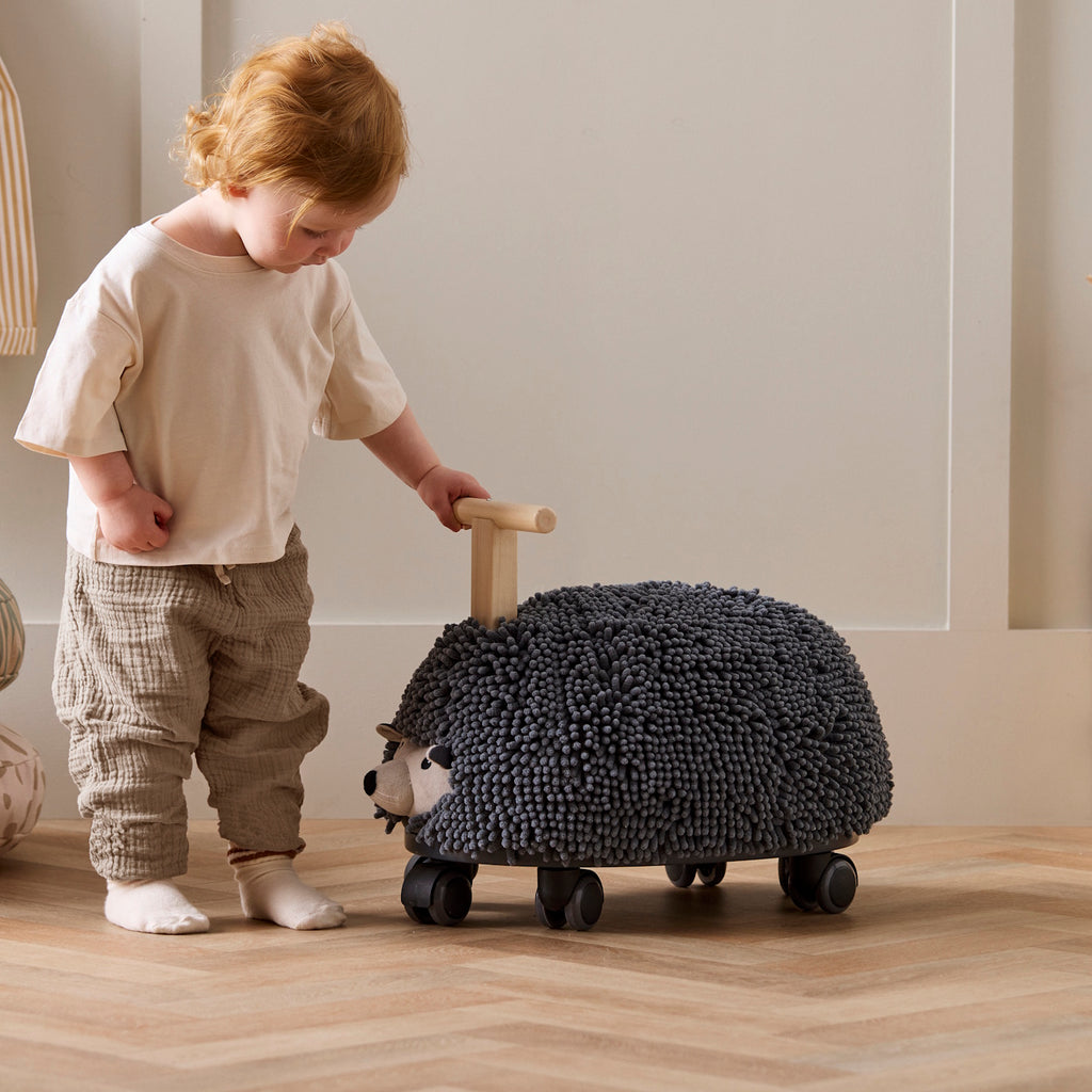 Toy hedgehog with a textured, dark gray surface resembling bristles, mounted on four wheels. It has a wooden handle for riding. Playful and whimsical.
