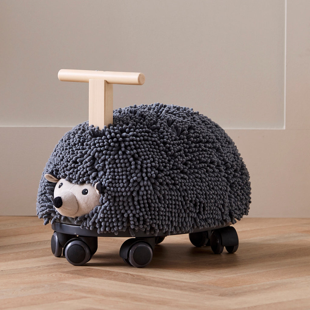 Toy hedgehog with a textured, dark gray surface resembling bristles, mounted on four wheels. It has a wooden handle for riding. Playful and whimsical.