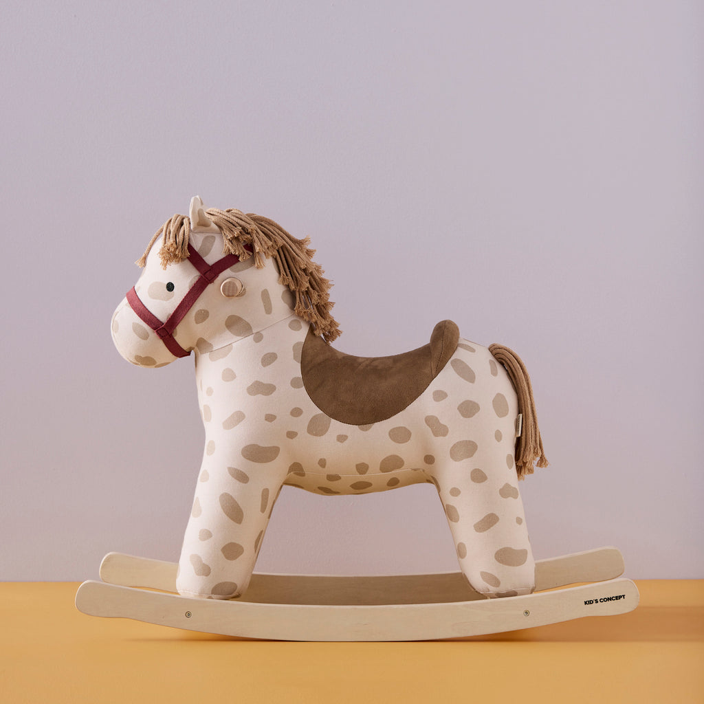 Plush rocking horse with a spotted beige pattern, red bridle, brown mane, and tail. It sits on a light wooden rocker against a soft purple backdrop.