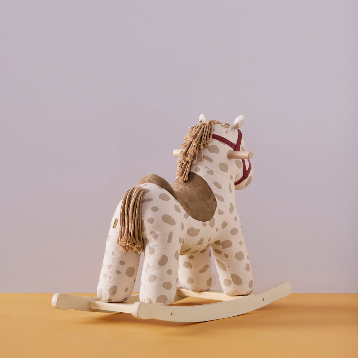 Plush beige spotted rocking horse toy with yarn mane, wooden base, and red bridle on a yellow surface.