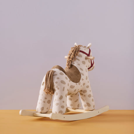 Plush beige spotted rocking horse toy with yarn mane, wooden base, and red bridle on a yellow surface.