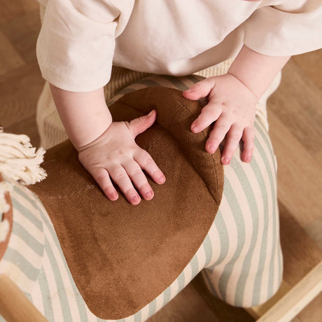 Close-up of a toddler's hands resting on the brown suede saddle of a striped rocking horse.