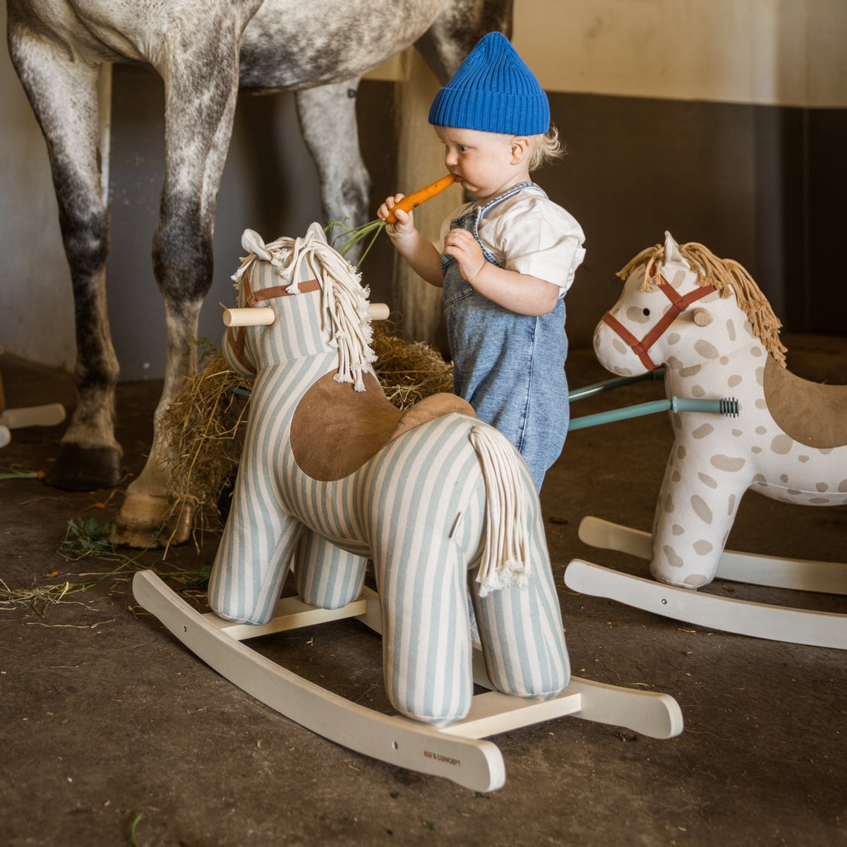 Toddler in a blue beanie feeding a carrot to a striped plush rocking horse in a stable.