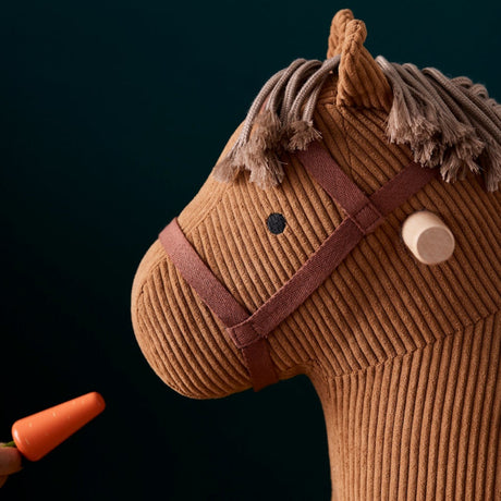 Close-up profile of a brown corduroy rocking horse head being offered a small toy carrot.