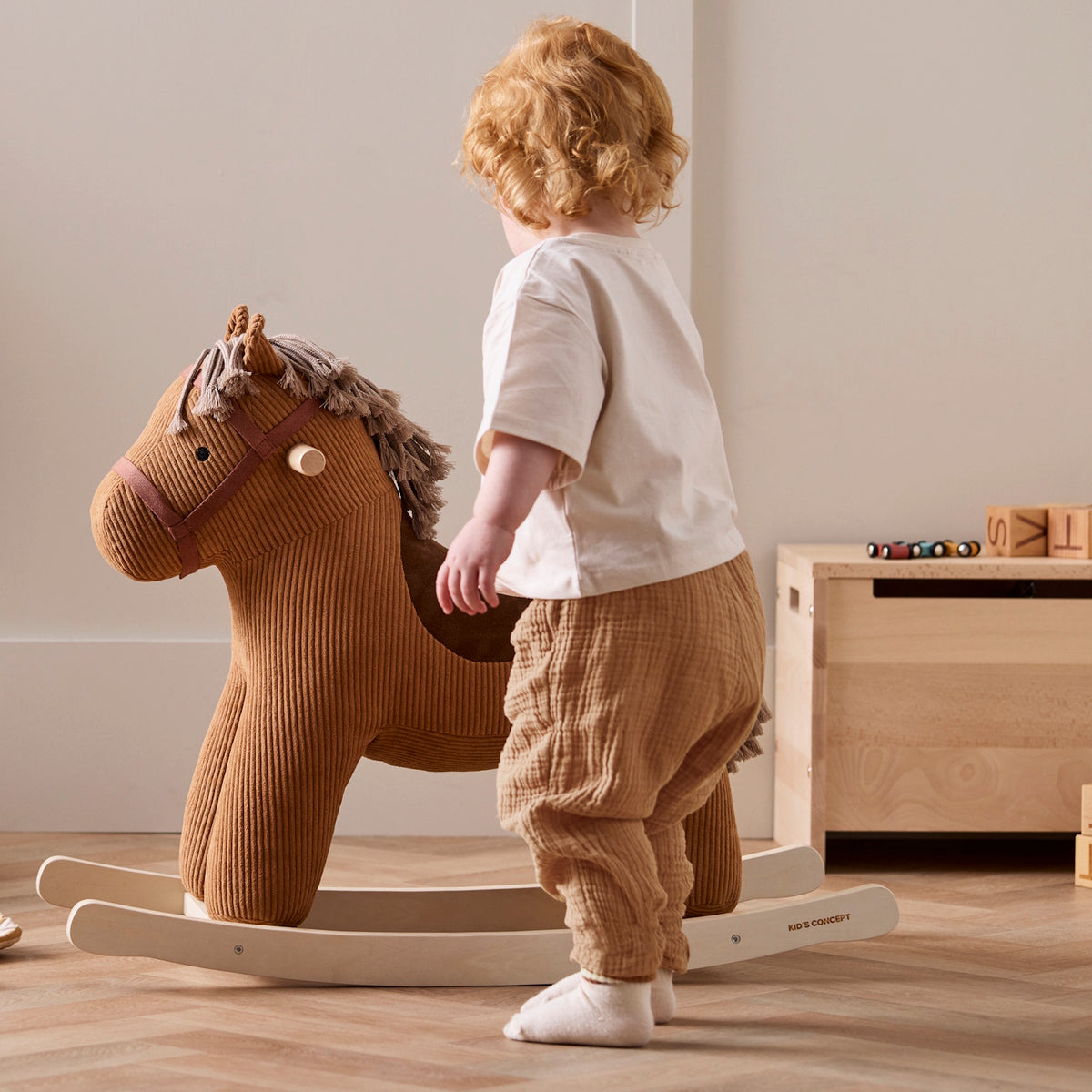 Toddler standing beside a plush brown corduroy rocking horse on wooden runners.