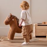 Toddler standing beside a plush brown corduroy rocking horse on wooden runners.