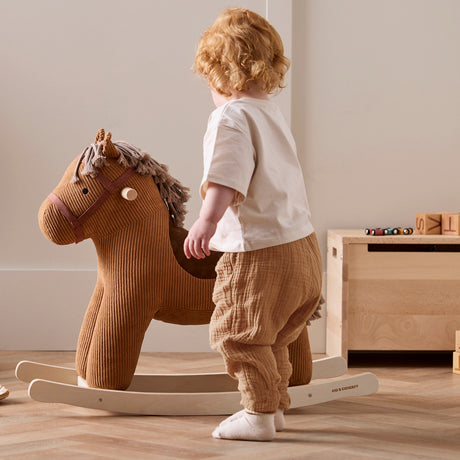 Toddler standing beside a plush brown corduroy rocking horse on wooden runners.