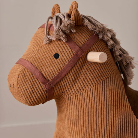 Close-up of a brown corduroy rocking horse toy with a fringe mane, bridle detail, and wooden handle.