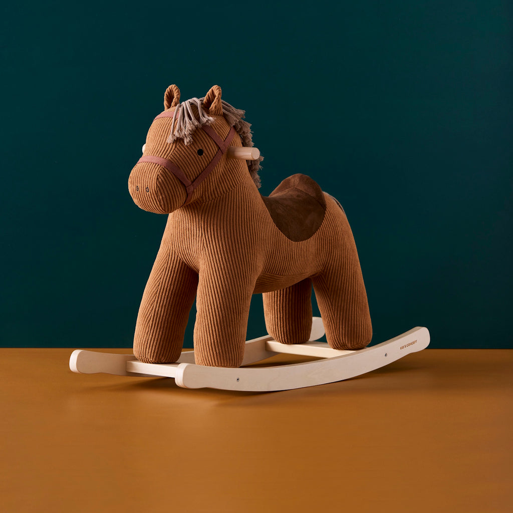 Plush rocking horse toy with a brown corduroy texture, wooden rocking base, and stitched details. The horse has a soft saddle and mane, evoking a cozy, playful feel.