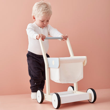 Toddler standing behind pushing a wooden toy shopping trolley with a fabric basket.