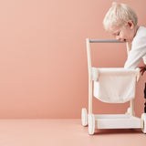 Toddler reaching into a wooden toy shopping trolley against a pink background.