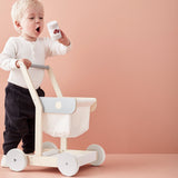 Toddler standing beside a wooden toy shopping trolley holding up a toy food can.