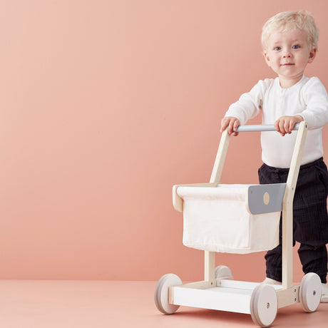 Toddler pushing a wooden toy shopping trolley against a pink backdrop.