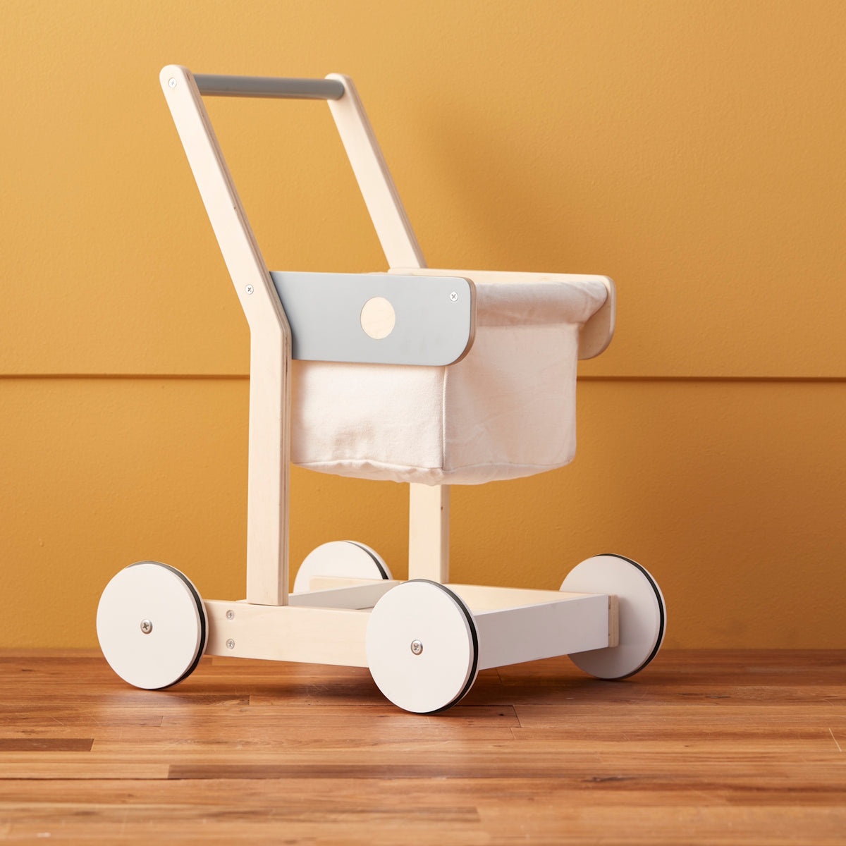 Wooden toy shopping trolley with a fabric basket set against a yellow wall and wood floor.