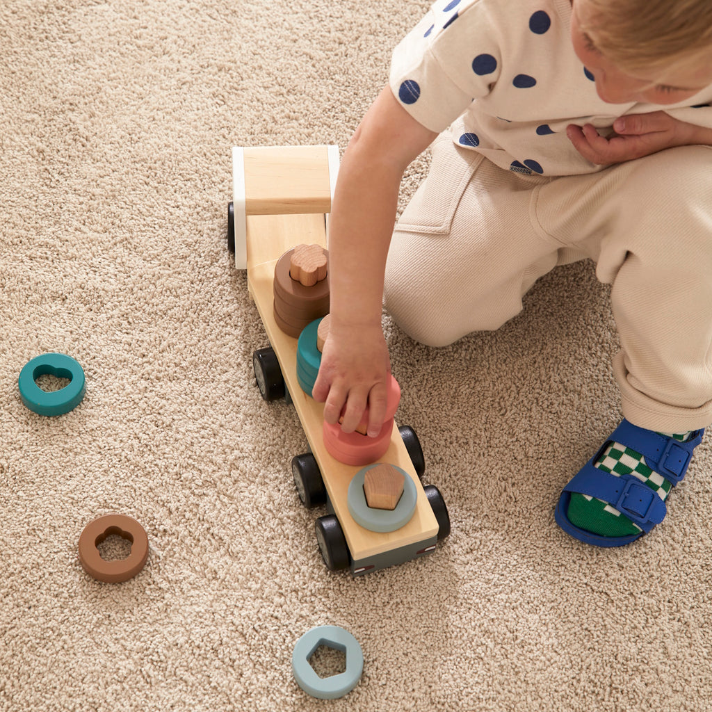 A wooden toy truck with a white cab and black wheels. The flatbed carries cylindrical wooden stacks in gray, pink, teal, and brown, creating a playful and organized look.