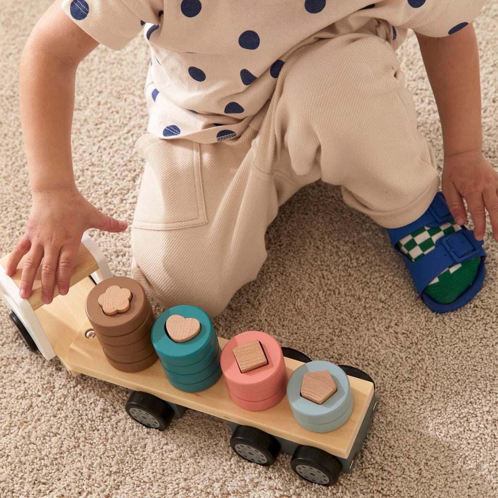 A wooden toy truck with a white cab and black wheels. The flatbed carries cylindrical wooden stacks in gray, pink, teal, and brown, creating a playful and organized look.