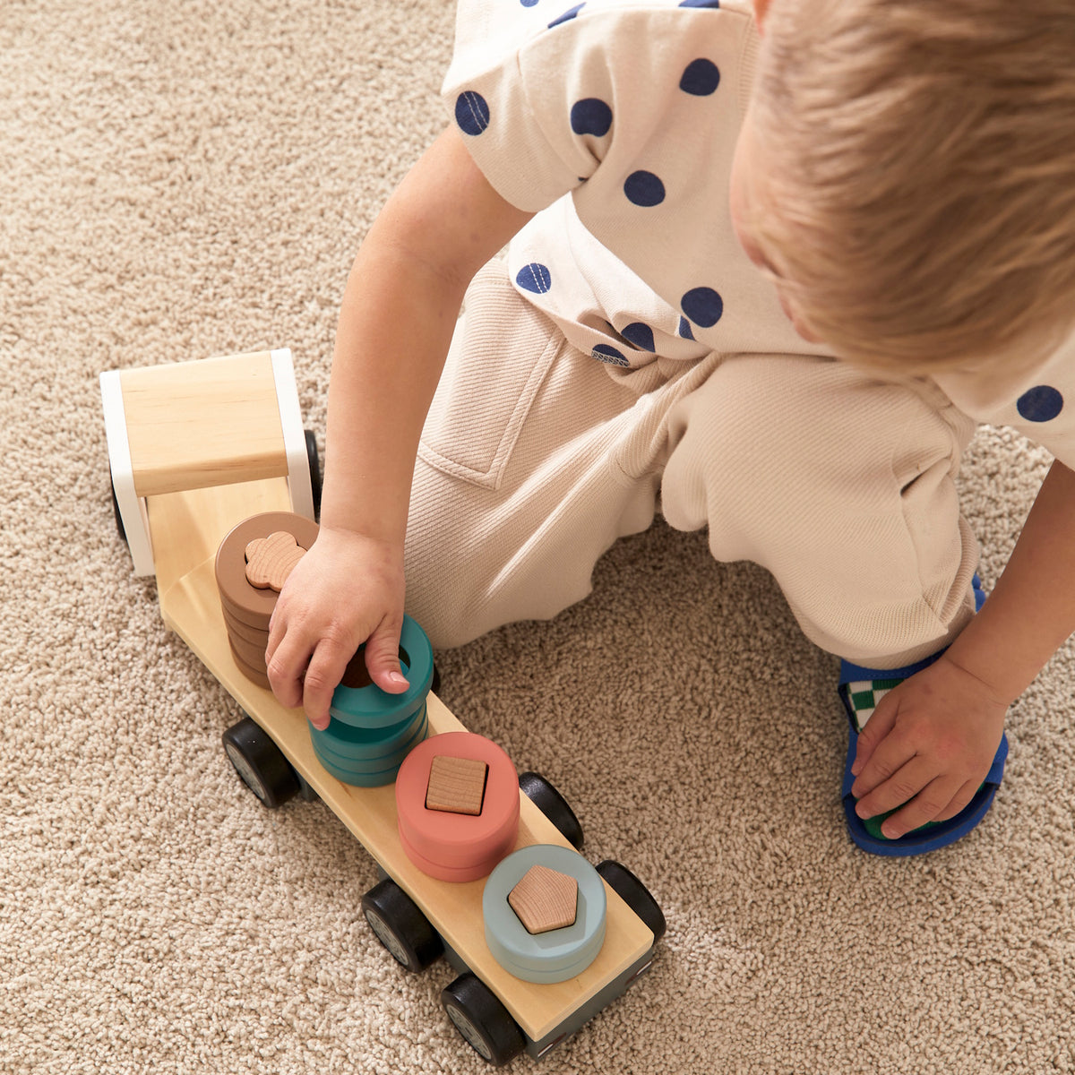 Child playing with a wooden toy truck stacking colorful shape-sorting rings on a beige carpet.