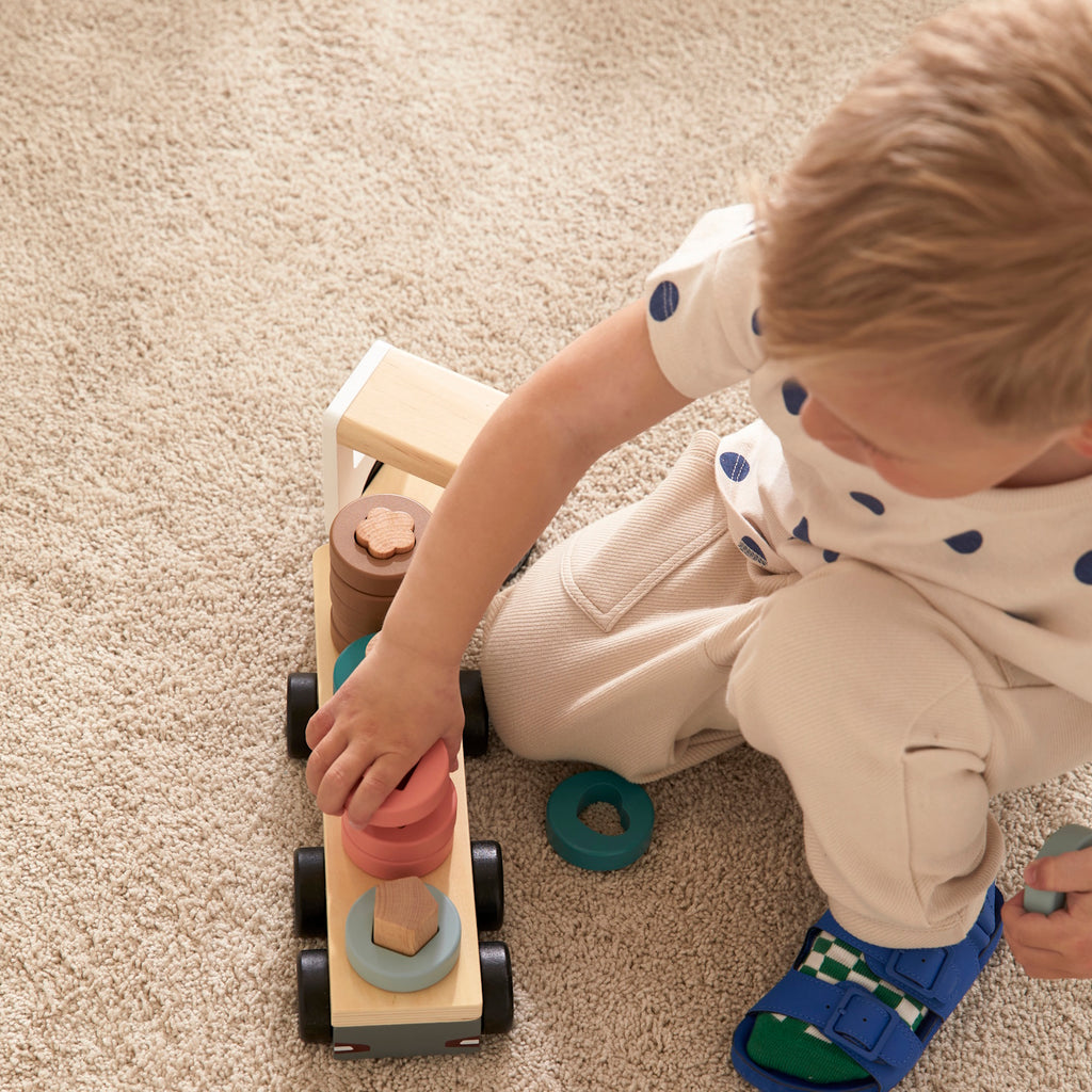 A wooden toy truck with a white cab and black wheels. The flatbed carries cylindrical wooden stacks in gray, pink, teal, and brown, creating a playful and organized look.
