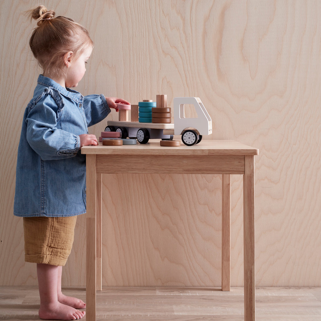 A wooden toy truck with a white cab and black wheels. The flatbed carries cylindrical wooden stacks in gray, pink, teal, and brown, creating a playful and organized look.