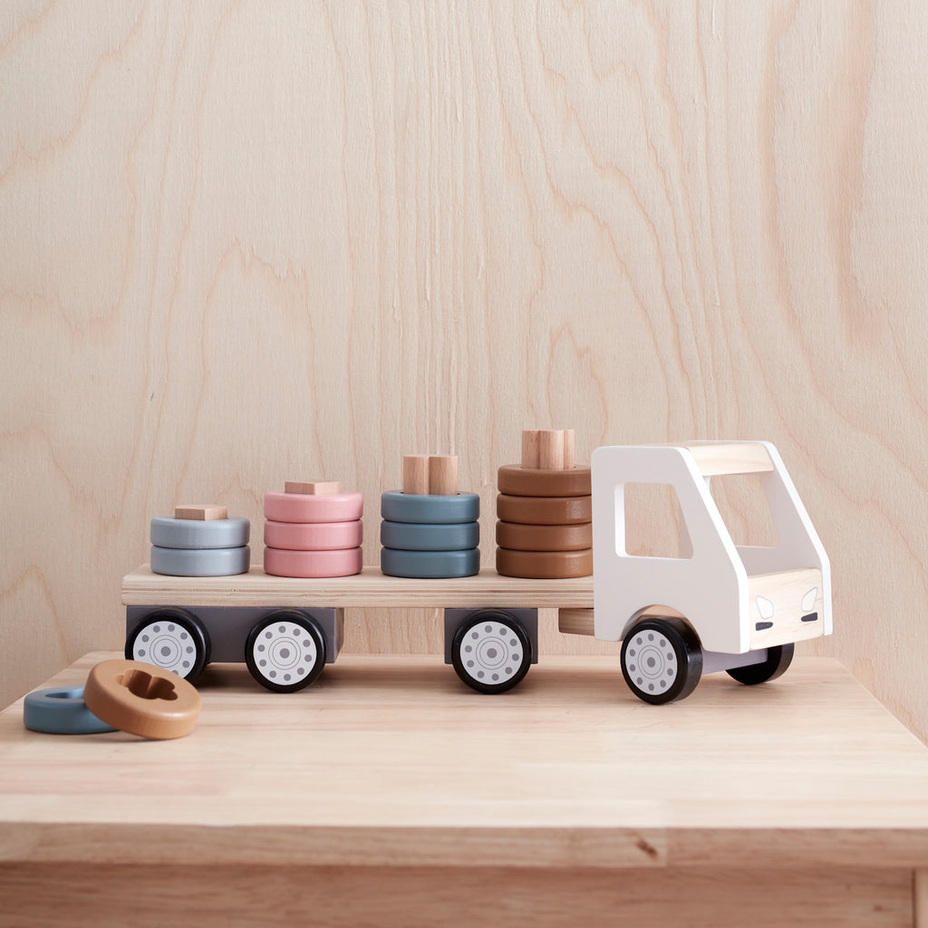 A wooden toy truck with a white cab and black wheels. The flatbed carries cylindrical wooden stacks in gray, pink, teal, and brown, creating a playful and organized look.