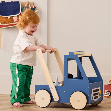 Toddler in green pants pushing a blue wooden toy walker truck across a room.