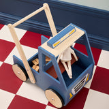 High-angle view of a blue wooden toy walker truck carrying a ragdoll and blocks on a checkered floor.