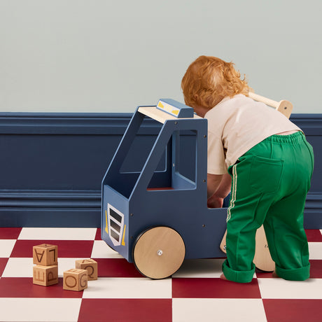 Toddler leaning over a blue wooden toy walker truck next to wooden alphabet blocks on a checkered floor.